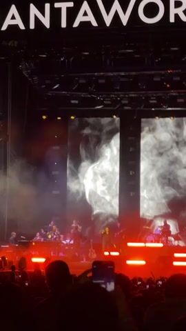 A live music performance is taking place on a large stage at night. The band, illuminated by orange stage lights, consists of a singer, keyboardist, guitarist, drummer, and several string musicians. Behind the band, three large screens display moving white graphics resembling smoke. The word "DANTAWO" is prominently displayed in large white letters above the stage. The crowd in the foreground appears as dark silhouettes. Some members of the audience are holding up their phones to record the performance.