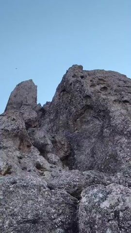 The video captures a low-angle shot of rocky formations against a clear blue sky. A small dark object, possibly a bird, is visible in the sky. The rocks are textured and weathered, with some appearing jagged and others more rounded. The lighting suggests it is daytime, possibly morning or late afternoon. The camera appears to be stationary, with slight vibrations.