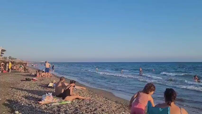 A beach scene under a clear blue sky shows many people enjoying the ocean. In the foreground, two individuals are seated on chairs facing the water, with a child standing between them. Further down the beach, several people are sunbathing on towels on the pebbly shore. The ocean is calm with small waves, and numerous people are swimming and playing in the water. The horizon is visible with a clear distinction between the blue sea and the lighter blue sky.