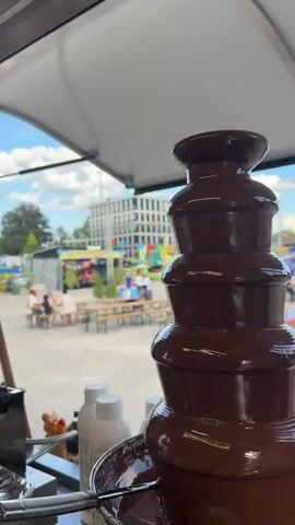 A close-up shot of a chocolate fountain is in the foreground, with a blurry outdoor scene in the background. The scene appears to be a festival or outdoor market with people milling about, picnic tables, and a stage with performers. There is a modern building in the distance, and the sky is partly cloudy. The overall impression is one of a lively and festive atmosphere.