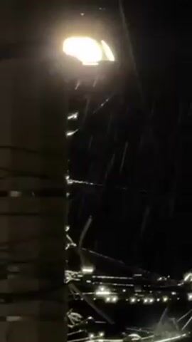 The video shows a night scene with rain falling under a street light. The street light is bright and casts a glow on the falling rain. There are many wires overhead, tangled and crisscrossing each other, silhouetted against the dark sky. A telephone pole is visible on the left side of the frame. The overall environment is dark and wet.