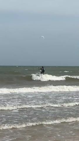 A kitesurfer rides a wave on the ocean under a cloudy sky. The kitesurfer is wearing a dark wetsuit and is holding onto the kite's bar. There is another kite in the distance, and more people can be seen in the water in the background. The waves are choppy, and there is white foam on the water. The beach is visible in the foreground, with sand and some seaweed.