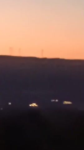 This is a video captured at dusk, featuring a blurred silhouette of a hilly landscape against a gradient orange sky. Several faint, indistinct lights are visible in the mid-ground, suggesting distant human settlements or infrastructure. On the crest of the hills, a row of tall, slender structures, possibly wind turbines, are silhouetted against the sky. The overall visual quality is low-resolution and out of focus, making precise identification difficult.