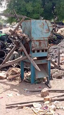 The video shows a rundown outdoor area filled with various objects. In the center stands an old, rusty machine painted in light blue. Debris and miscellaneous items surround it, including tires, buckets, bags, and pieces of wood. A large pile of garbage, consisting mostly of white plastic bottles, is visible in the background, partly covered by a tan sheet. A large green tree stands in the upper background. The ground is dirt and littered with trash. The overall impression is of a cluttered and perhaps neglected workspace.