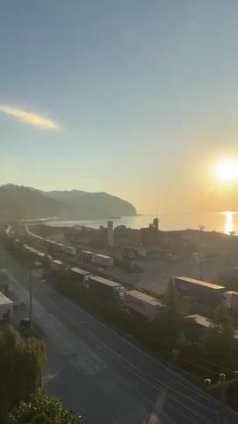 The video captures a sunset over a coastal industrial area. A bright sun hangs low in the sky, casting a warm glow and a reflection on the calm sea. In the foreground, a highway runs parallel to the coast, with numerous semi-trucks parked along its side and on a dirt lot. Some of the trucks are white with red accents, while others are entirely white or have darker colored trailers. A few trees, including a weeping willow, are visible along the edge of the highway. In the background, industrial buildings, including a tall, square structure and what appears to be a silo, are scattered throughout the landscape. Mountains are visible in the far distance, partially obscured by haze. The overall scene suggests a working port or industrial zone bathed in the golden light of dusk.