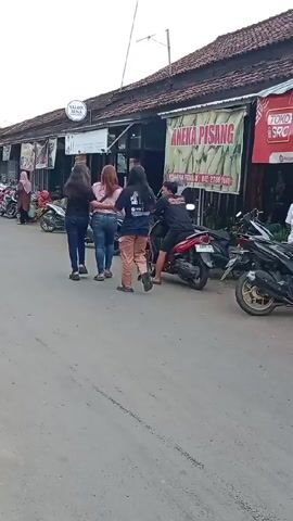 The video shows a street in what appears to be a small town. There are several parked motorcycles on the side of the road, and people walking along the sidewalk in front of a row of shops. One woman is riding a motorcycle away from the camera. The shops have signs that read "SALON IRNA," "ANEKA PISANG," and "TOKO SRC." The video is taken from a stationary position.