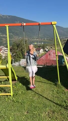 A young woman with blonde hair and fair skin is sitting on a swing set, which is set in a grassy area. She is wearing a white dress and a denim jacket. The swing set is green and orange, and there is a slide attached to it. In the background, there are houses and mountains. The sky is blue and clear. The woman is smiling and looking up at the sky. She appears to be enjoying herself. The video is a short clip, lasting only a few seconds. The camera is positioned in front of the swing set, and it captures the woman as she swings back and forth. The lighting is bright, suggesting that it is daytime.