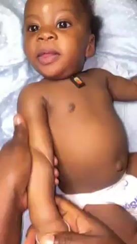 A baby is laying on its back on a blue and white polka-dotted surface, wearing only a diaper and a small, rectangular pendant or tag around the neck. An adult is using both hands to massage the baby's left arm, starting from the wrist and moving up towards the shoulder. The baby is looking directly at the camera. The lighting is well-lit and the camera angle is slightly above the baby, looking down.