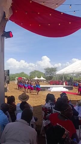 The video shows a cultural performance taking place outdoors.  A group of men dressed in traditional attire, consisting of blue and pink skirts and carrying sticks, is dancing in a field. They are performing in front of a wedding party, who are gathered near a stage. Spectators are seated under a tent with red draped fabric and string lights. The sky is blue with scattered clouds.