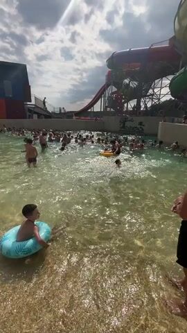 The video shows a crowded wave pool at a water park. In the foreground, a young boy wearing a blue swim ring with a palm leaf pattern is facing away from the camera, partially submerged in the water. Several other people are visible in the water, some with swim rings, others swimming or standing. In the background, a large water slide structure with red, purple, and green tubes is prominent. People can be seen on the stairs leading up to the slides. The water is clear and has small waves. The sky is cloudy with patches of sunlight.