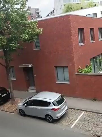 A camera pans across a street with parked cars and brick buildings. The street is paved with cobblestones. A silver hatchback is parked on the left side of the street. The building to the right is made of red brick. The camera then pans to the right, revealing another red brick building with windows and an awning. A white car with a black roof is parked on the right side of the street.