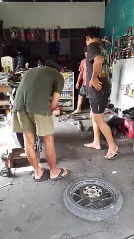 Two men are in a cluttered garage. One man, wearing a green t-shirt and khaki shorts, is bent over and appears to be working on a motorcycle. He is wearing sandals. A second man, wearing a red t-shirt and patterned shorts, stands a few feet away, observing. He is wearing sandals and a baseball cap. A motorcycle wheel lies on the floor in front of them. Shelves filled with various items are visible in the background.