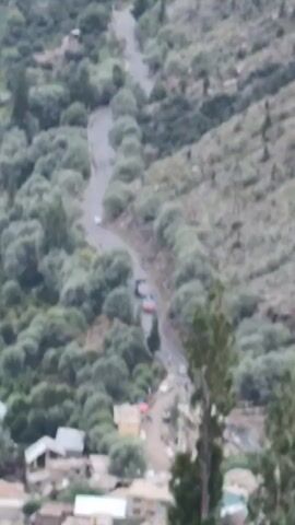 The video begins with a high-angle shot of a winding road on a steep, tree-covered mountainside. Several cars are visible on the road, some traveling uphill and others downhill. The road passes by a small village with houses and shops. As the video progresses, the camera pans out to reveal a wider view of the valley. The winding road continues through the valley, which is dotted with more villages and patches of farmland. A river flows alongside the road in the distance. The video ends with a panoramic view of the valley, with the river snaking its way through the landscape and the mountains rising steeply on either side. The sky is overcast with some clouds visible.