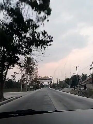 The video shows a road with an archway ahead, seemingly marking an entrance to a town or region. The sky is overcast, suggesting it's either early morning or late afternoon. The road is paved and appears to be a two-lane highway. There are trees and vegetation on either side of the road, including a palm tree on the left. A motorcycle is visible on the right side of the road, moving away from the camera. The archway is ornate, with decorative elements and text, possibly in Arabic script, above an inscription in Latin script that reads "SELAMAT DATANG DI KABUPATEN LAMPUNG UTARA". This translates to "Welcome to North Lampung Regency". There are utility poles with wires visible along the road. The overall scene depicts a rural or semi-rural setting with a clear visual indicator of a territorial boundary. The camera appears to be mounted inside a vehicle, as evidenced by the dashboard visible at the bottom of the frame. The video is shot from a moving perspective, capturing the approach to the archway.