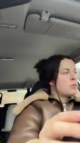 A young woman is sitting in the driver's seat of a car. She's wearing a brown leather jacket with a fluffy, light-colored collar. She's looking around, pursing her lips slightly, and appears to be paying attention to her surroundings while driving. The interior of the car is visible, including the dashboard, the rearview mirror, and the headrest of the driver's seat. Through the car window, we can see a glimpse of the outside environment, including trees and possibly a road. Music plays in the background.