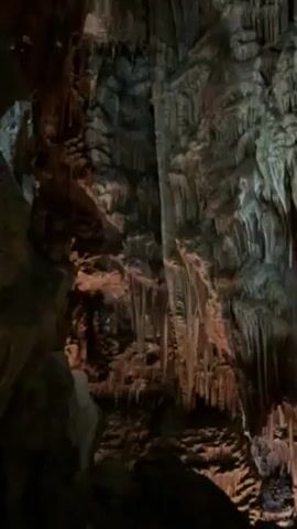 The video shows the inside of a cavern. The cavern walls are covered in formations of rock, mostly stalactites. The rock is a dark gray color, with some parts appearing brown. The lighting is dim and uneven, highlighting the textures of the formations. The camera is stationary.