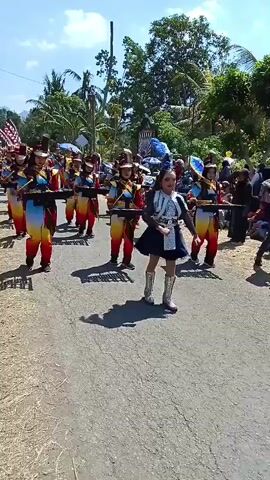 A marching band composed of young women, all wearing matching uniforms consisting of hats, jackets, and pants with red, yellow, and blue gradients, are marching down a paved road. They are carrying instruments that resemble keyboard instruments with black keys. The band members are walking in formation, with one individual in the foreground, wearing a similar uniform but with a blue skirt and silver boots, appearing to lead them. The scene is outdoors on a sunny day, with trees and vegetation visible in the background. There are also spectators lining the sides of the road.
