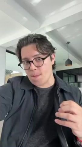 A young man with dark hair and glasses is standing indoors, adjusting his hair. He's wearing a gray sweater and a dark-colored jacket. He runs his fingers through his hair several times, then adjusts the collar of his jacket. The background shows a modern interior with a window, a light fixture, and a ceiling with recessed lighting. He appears to be looking directly at the camera throughout the video.