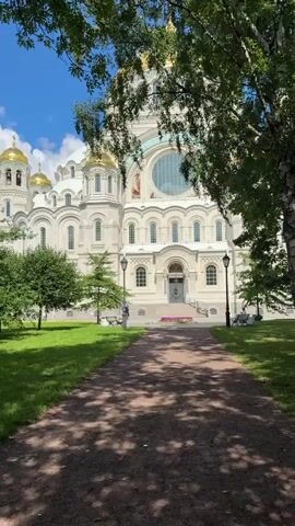 The video shows a medium shot of a grand white cathedral with golden domes. The cathedral is seen from across a green lawn and a tree-lined path. The path is dappled with sunlight and shadows from the trees. In the foreground, there are trees with green leaves, and in the background, there is a bright blue sky with scattered clouds. The camera zooms in slightly on the cathedral, revealing intricate architectural details, including arched windows, decorative carvings, and mosaics above a large circular window. There are two lampposts on either side of the path leading to the cathedral entrance. Some people can be seen walking on the lawn to the left of the cathedral. The overall scene is serene and picturesque, highlighting the beauty of the cathedral and its surroundings on a sunny day.