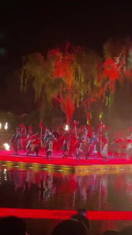 At night, people watch a fiery outdoor performance on a stage floating in water. Performers in red and black clothing wield swords and sticks lit with fire. The performance is near trees and includes a large burst of fire. The audience records with phones.