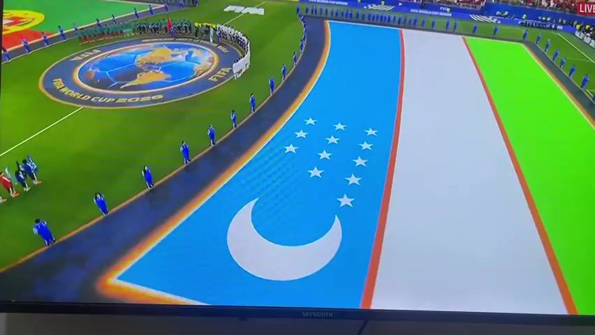 An aerial view of a soccer stadium shows a large flag spread out on the grass, resembling the flag of Uzbekistan. In the background, players in blue uniforms stand in a line along the edge of the field, while other players in red and green uniforms are seen on the field. A circular logo with a globe is visible in the center of the field. The stadium is filled with spectators in red shirts. The camera then zooms in on a close-up of a young man's face, who is looking upwards with his mouth open. He appears to be a soccer player, possibly singing the national anthem, given the context of the stadium setting and the presence of flags and teams. The screen also shows "ZOR TV LIVE" and "FIFA WORLD CUP 2020" in the corner.