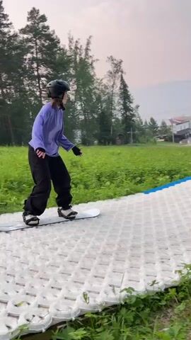 The video shows a person wearing a helmet and purple long-sleeved shirt, black pants, and gloves, snowboarding on a textured ramp. The person begins on a white, textured surface, then moves onto a black, cylindrical obstacle, and attempts to transition to a blue surface. The snowboarder falls onto the blue surface. The environment is grassy with trees and buildings in the background.