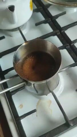 A close-up, overhead shot shows a metal cezve filled with coffee grounds and water heating on a gas stove. The stove has a white surface with a black grate. The cezve is positioned over a lit burner, and small bubbles are beginning to form on the surface of the coffee. In the background, a white teapot with yellow floral accents can be seen. A small amount of liquid has spilled onto the stove surface near the cezve. The scene is lit from above, creating a slight shadow.