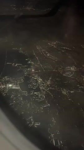 The video shows a night cityscape from an airplane window. The city lights appear as scattered points and lines of yellow and white. The view is slightly obscured by condensation or fog on the window. The airplane wing is faintly visible in the upper corner of the frame.