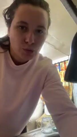A woman with her hair pulled back in a bun is shown from the chest up washing a white plate with blue and pink designs on it. She is wearing a long-sleeved light pink shirt. Behind her is a window with a curtain featuring a bright yellow and orange geometric pattern. A black object, possibly a kitchen fixture, is visible to her right. The lighting is bright and somewhat harsh, giving the impression of being indoors under artificial light. She has a neutral expression as she scrubs the plate.
