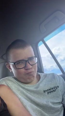 This is a close-up, waist-up, selfie-style video of a young person wearing glasses and a gray t-shirt with the words "knowledge power" printed on it. The person is sitting in the passenger seat of a car, with the sun shining through the window behind them. The person is smiling at the camera. The background is slightly blurred, but the sky and clouds are visible through the window.