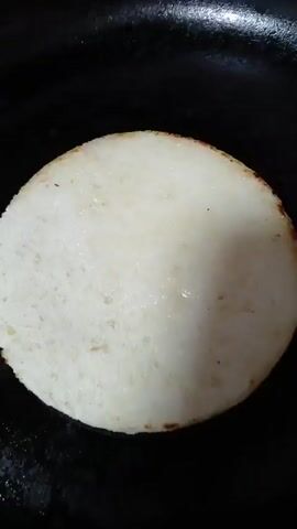 A close-up shot shows a circular, white batter being cooked in a black pan on a stovetop. The batter is evenly spread and appears to be rising slightly, with some small bubbles forming on its surface. The pan is heated by a gas stove, indicated by the visible burner elements beneath it. The lighting is somewhat dim, highlighting the cooking batter as the primary focus. The camera remains stationary throughout the short clip, capturing the cooking process from a top-down perspective.