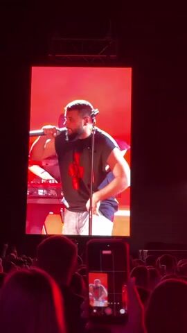 A concert is taking place, with a large screen displaying a close-up of a man singing into a microphone. The screen is illuminated with a bright red hue. He is wearing a sleeveless t-shirt with a graphic design and white pants. Behind him on the screen, another person wearing glasses and a black shirt can be seen. In front of the screen, a crowd of people is visible, many holding up their phones to record. A phone in the foreground is actively recording the screen, showing a smaller version of the performance. The stage is also lit with red lights, and other musicians can be seen on stage, one playing a guitar and another at a keyboard.