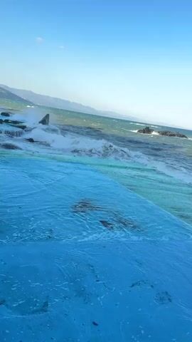 A videocam pans across a rough sea under a clear blue sky. The ocean is choppy and waves crash against dark rocks jutting out of the water. In the foreground, a flat, blue-painted surface slopes down towards the sea. The surface appears to be a walkway or a swimming pool area, with the blue paint extending into the shallow water. The water is clear and reveals a submerged ladder structure in the distance. The overall scene depicts a coastal environment with rough seas and clear skies.