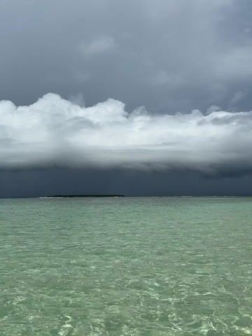 The scene is a still shot of a calm, clear, light green ocean under a stormy sky. A small, dark green island is visible in the distance, under a thick layer of dark storm clouds. The sky above the storm clouds is lighter and gray. The water's surface is clear, revealing subtle patterns and textures. No people or other objects are visible in the foreground or background. The focus is on the contrast between the tranquil water and the looming storm clouds.