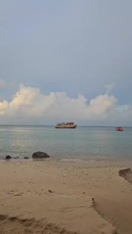 The video shows a beach with calm, turquoise water and a cloudy sky. In the distance, there are two boats. The closest boat is yellow with a red roof, and the other boat is red. The beach is made of sand and there are a few rocks visible in the water near the shore. The video appears to be filmed from a stationary position on the beach, looking out towards the sea.