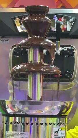 A close-up shot shows a chocolate fountain in operation. Melted chocolate cascades down three tiers of the fountain, pooling in a stainless steel basin at the bottom. The machine is mostly silver, with some colorful graphics visible on the top panel, including images of ice cream cones. The base of the fountain has decorative cutouts resembling butterflies and warning labels. The lighting appears to be overhead and casts reflections on the shiny surfaces. No people are visible.