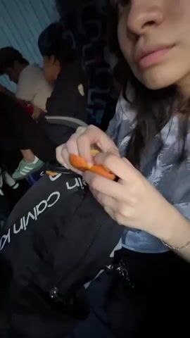 A person is seen peeling a small orange fruit, likely a mandarin or clementine, inside a bus. The person is wearing a grey and white tie-dye long-sleeved shirt, with a black Calvin Klein backpack resting on their lap. In the background, other passengers are visible; one person wears light green Adidas sneakers, and another is looking at a smartphone. The bus interior features blue seats with a patterned fabric.
