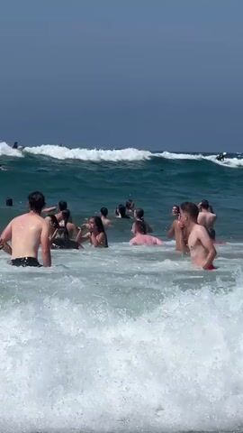 This is a video clip of a group of people swimming and playing in the ocean on a sunny day. The water is a vibrant turquoise green, with waves breaking and creating white foam. In the foreground, several individuals are wading in the shallow water, while further back, a larger group is clustered together, some appearing to be talking or splashing each other. In the background, a wave is cresting, and a few people are visible on surfboards further out at sea. The sky is a clear, deep blue. The overall atmosphere appears to be one of enjoyment and recreation on a warm, bright day at the beach. The location is implied to be San Sebastián, Spain. There is no visible text in the clip.