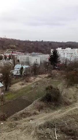 The video shows a view of a cityscape under an overcast sky. In the foreground is a vacant lot with dirt paths and sparse vegetation, including some small trees without leaves. In the middle ground, there are a few small buildings and gardens. In the background, there are several white apartment buildings, some with colored accents. On the horizon, there is a line of trees and a few taller structures. The camera pans slightly to the right during the video, revealing more of the cityscape.
