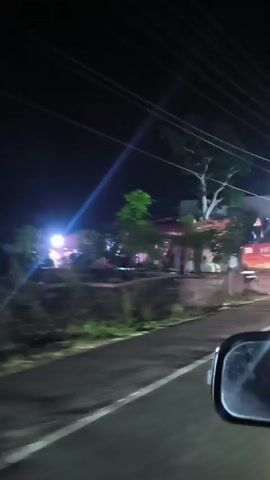 The video shows a nighttime scene from a moving vehicle. The camera pans across a road and then focuses on a large, red Hitachi excavator parked on the side of the road. The excavator is massive, with large tracks and a prominent boom. There are trees and some indistinct structures behind the excavator. A few lights are visible, illuminating the scene. A person is seen walking in the foreground, and another person is standing near the excavator. The road appears to be paved, and there are utility lines overhead. The overall impression is of an active construction site or a recently completed one.
