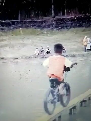 The video shows a child on a bicycle riding on a narrow wooden plank over water. The child is wearing an orange shirt and green shorts. In the background, there are people watching and what appears to be a race or event taking place. There are trees and a building in the distance. The video is grainy and appears to be of low quality. The child is focused on balancing and riding the bicycle across the plank.