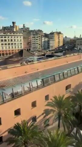 The video shows an urban scene with buildings, palm trees, and structures with glass roofs. The buildings are various colors and designs, with some having towers. There are roads and possibly train tracks visible between the buildings. The sky is blue with some clouds. There are also glimpses of industrial elements like cranes in the distance. People's voices can be heard in the background. The perspective is from an elevated position, possibly a window or balcony.