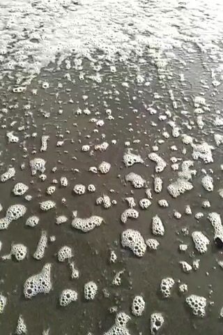 The video shows a beach with dark sand and small waves rolling in. The tide is low, and there is a lot of sea foam on the sand. The sky is cloudy with patches of blue.