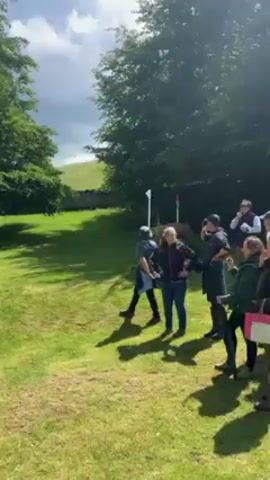 Here is a detailed description of the video:

The video captures a scenic view of an equestrian event taking place on a sprawling, grassy field on a sunny day. At the start of the video, a group of people are seen standing on the grass, some with their backs to the camera. As the camera pans to the right, a couple of women are seen pointing towards a small lake. 

The lake is decorated with multiple obstacles, including what appears to be a large, wooden log laid across the water with flowers planted on it. There's also a small evergreen tree on the edge of the lake. The far side of the lake features a larger structure with a white tent visible in the background, and yellow signs. There are other smaller green obstacles with white and red flags in the lake.

The area is surrounded by lush green trees and foliage. The sky is bright with scattered clouds, casting shadows on the grassy field. Overall, the scene captures a vibrant and engaging outdoor event.