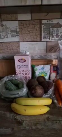 The video shows a collection of groceries laid out on a wooden surface. In the foreground, two yellow bananas are visible. Behind them, there are several beets in a plastic bag, and to the left, a bag of cucumbers. To the right of the beets, a bunch of carrots is arranged. In the background, a carton of milk with Russian labeling indicating "MILK 3.2%" stands prominently. Next to the milk is a bag of what appears to be flour or a similar dry good, also with Russian labeling. Further to the right, a box of cereal or pasta and a large plastic bottle of water are present. The backdrop is a tiled wall with a pattern resembling brick and a cityscape with houses and trees. The lighting is moderate, and the camera angle is slightly from above, looking down at the groceries.