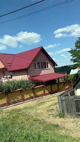 This is a landscape video shot from a moving vehicle, likely a car. The video pans across a rural scene with a clear blue sky dotted with white clouds. In the foreground, there is green grass and a partially visible power pole. Further back, a house with a red roof and light-colored walls is visible, partially obscured by green bushes and a fence. A metal fence with brick pillars lines the property. Beyond the house, more trees and vegetation can be seen, along with other houses in the distance. A traditional well with a roof is also visible in the middle ground. The video continues to pan, revealing a cornfield and another house with a weathered wooden fence and a corrugated roof. The overall impression is that of a sunny day in a village or countryside setting.