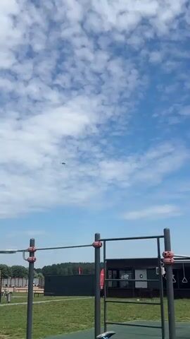 A drone is filming a small airplane flying in the sky. The drone is filming from a low angle, looking up at the sky. In the foreground, there are some horizontal bars of a playground. In the background, there is a building and trees. The sky is blue with some white clouds. The airplane is white with some red and blue markings. The airplane is flying from left to right.