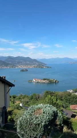 The video shows a panoramic view of a large lake with islands and distant mountains under a clear blue sky with some scattered clouds. In the foreground, there is a residential building with a satellite dish and lush green vegetation including cypress trees. The middle ground features a picturesque island dotted with buildings with terracotta roofs and a small harbor with boats. Another, smaller island is visible further in the distance, also with buildings. The background reveals a coastline with a town nestled against rolling hills and the majestic outline of mountains fading into the horizon. The water of the lake is a deep blue and appears calm, with a few small boats scattered across its surface. The overall impression is one of a serene and beautiful landscape, likely a tourist destination.