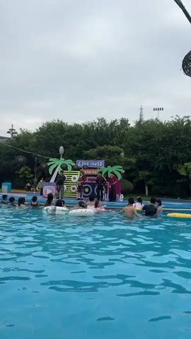 The video shows a pool party event. In the foreground, the blue water of a swimming pool is visible, with several people of varying ages swimming and floating on inflatable rings. In the background, a stage area is set up with a backdrop featuring palm trees and the words "LIVE HOUSE WATER BOYS." Four performers are on the stage, dressed in casual attire. Two of the performers appear to be male, wearing jackets and pants, while another is in a plaid shirt and shorts. The fourth performer is in a red outfit. Several inflatable rings are placed on the stage behind the performers. Trees and a cloudy sky are visible behind the stage. The overall atmosphere appears to be a festive outdoor event.