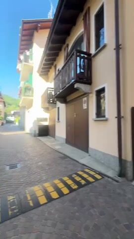 The video shows a young woman walking down a narrow street in what appears to be a European town. The street is paved with cobblestones and there are buildings on both sides. She is wearing a beige coat and a Burberry scarf. She is holding the camera up to film herself. Two people are walking with her, a man in black and another, younger, man wearing a hoodie that reads "BURBERRY." The sky is blue and the sun is shining.